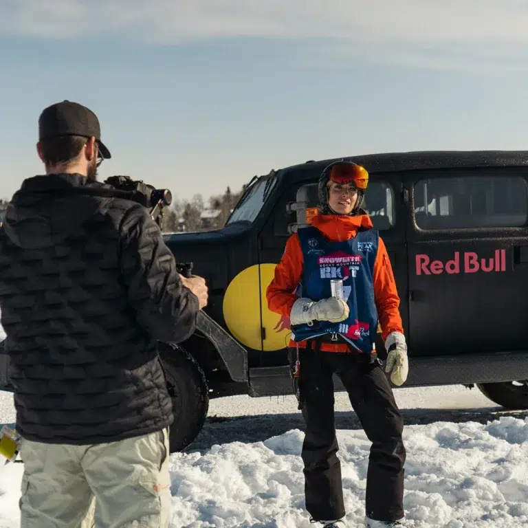 A snowboarder poses for the camera in front of an SUV for an event video.