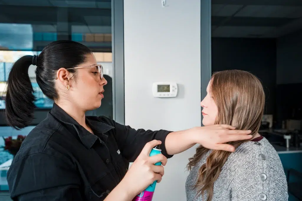 A hair and makeup artist applies hairspray to a client before a video shoot.